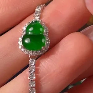 A close-up of a hand elegantly holding a Green Jade Gourd Bracelet with Silver Inlay, which showcases a smooth, green gourd-shaped jade centerpiece. The jade is encircled by sparkling stones set in a delicate silver frame, and the bracelet's band is adorned with similarly brilliant stones, creating a cohesive and luxurious piece of jewelry.