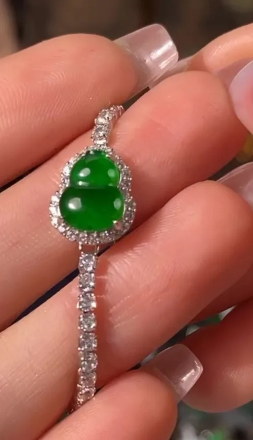 A close-up of a hand elegantly holding a Green Jade Gourd Bracelet with Silver Inlay, which showcases a smooth, green gourd-shaped jade centerpiece. The jade is encircled by sparkling stones set in a delicate silver frame, and the bracelet's band is adorned with similarly brilliant stones, creating a cohesive and luxurious piece of jewelry.