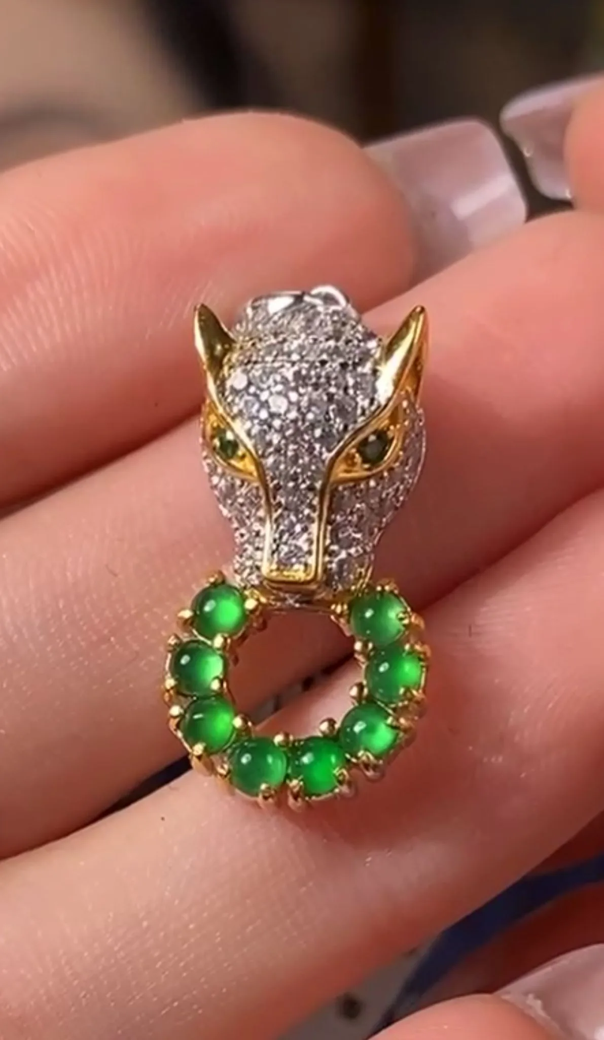 A hand delicately holds a Green Jade Pendant with Silver Inlay, showcasing a detailed fox head design with sparkling stones and golden accents. The fox's neck is adorned with a circle of luminous green jade beads set in a gold-tone base, creating an aura of elegance and mystery. The intricate craftsmanship and vivid colors of the pendant make it a captivating accessory.