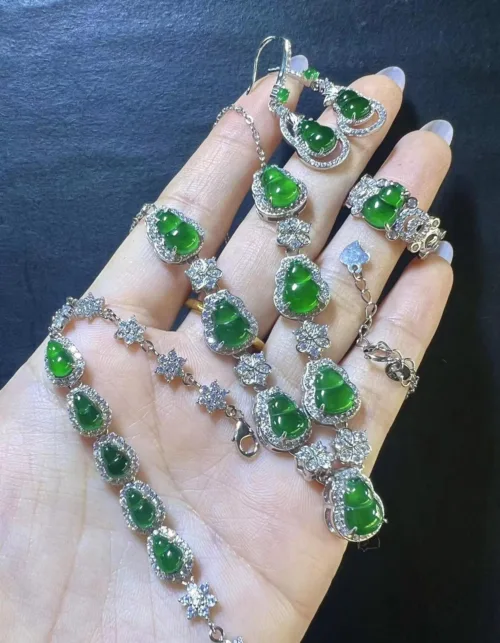 The image captures a hand holding a luxurious Jadeite Gourd Necklace. The necklace features multiple gourd-shaped jadeite charms with a deep green luster, each surrounded by a halo of twinkling stones set in silver. The charms are connected by a silver chain with additional star-shaped silver and stone accents, creating a continuous flow of elegance and harmony.