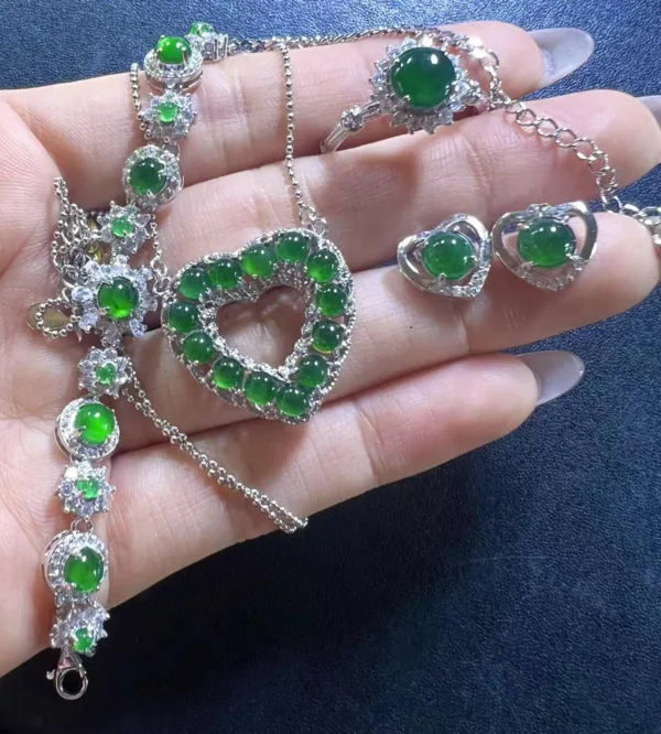 A hand holds a complete Jadeite Heart-Shaped Set against a dark blue fabric backdrop. The set includes a necklace with a central heart-shaped pendant surrounded by smaller jadeite hearts, a matching bracelet with linked heart gems, and heart-shaped stud earrings. Each jadeite heart glows with a vibrant green hue, elegantly framed by sparkling silver details, showcasing the set's cohesive and lovingly crafted design.