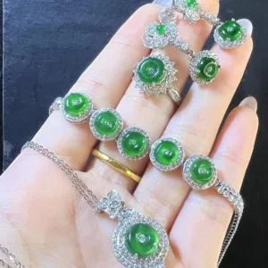 In the image, a hand is gently holding a Jadeite Peace Buckle Necklace, displaying its centerpiece—a lush green jadeite peace buckle surrounded by a glittering halo of silver and stones. Additional smaller jadeite pieces and silver details embellish the necklace chain, creating an elegant and cohesive piece that exudes tranquility and grace.