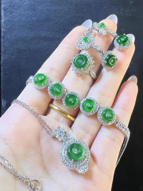 In the image, a hand is gently holding a Jadeite Peace Buckle Necklace, displaying its centerpiece—a lush green jadeite peace buckle surrounded by a glittering halo of silver and stones. Additional smaller jadeite pieces and silver details embellish the necklace chain, creating an elegant and cohesive piece that exudes tranquility and grace.