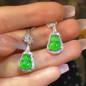 The image shows a pair of Jadeite Sun Green Gourd Earrings held elegantly between fingers. The earrings each feature a bright green jadeite carved into a gourd shape, symbolizing good luck, encased in a sparkling silver inlay with small, glittering stones accentuating the outline. The earrings dangle gracefully, reflecting the light and showcasing the vivid, translucent quality of the jade.