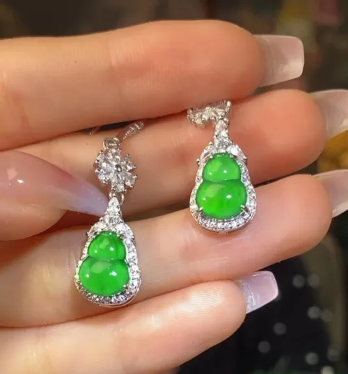 The image shows a pair of Jadeite Sun Green Gourd Earrings held elegantly between fingers. The earrings each feature a bright green jadeite carved into a gourd shape, symbolizing good luck, encased in a sparkling silver inlay with small, glittering stones accentuating the outline. The earrings dangle gracefully, reflecting the light and showcasing the vivid, translucent quality of the jade.