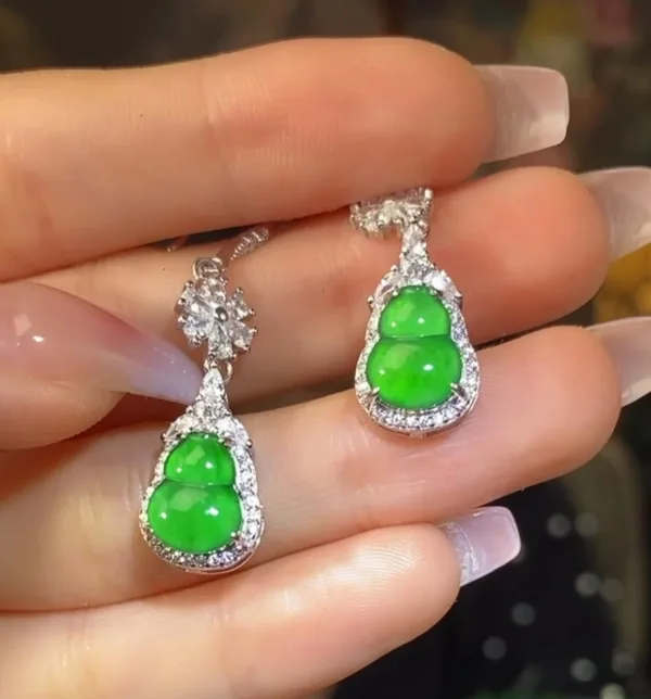 The image shows a pair of Jadeite Sun Green Gourd Earrings held elegantly between fingers. The earrings each feature a bright green jadeite carved into a gourd shape, symbolizing good luck, encased in a sparkling silver inlay with small, glittering stones accentuating the outline. The earrings dangle gracefully, reflecting the light and showcasing the vivid, translucent quality of the jade.