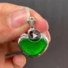 A hand holding a "Ruyi Pendant (Gold Inlaid)" featuring a heart-shaped vibrant green gemstone with visible inclusions, set in a silver mount with gold inlay and intricate detailing. The pendant is suspended from a silver loop encrusted with small diamonds.