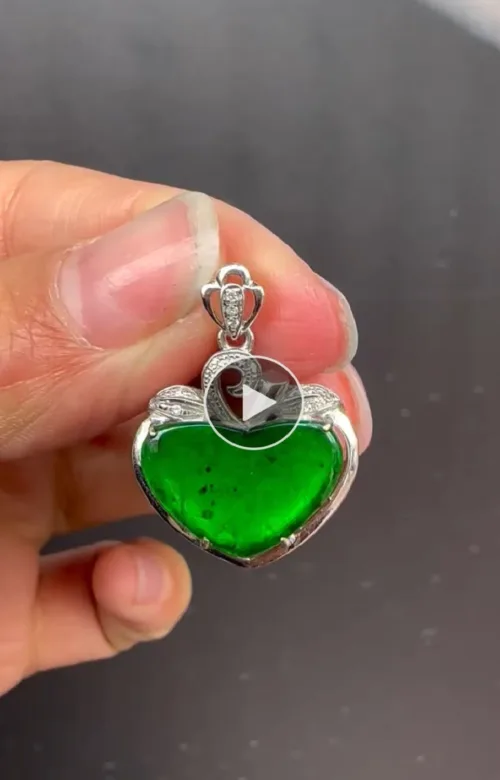 A hand holding a "Ruyi Pendant (Gold Inlaid)" featuring a heart-shaped vibrant green gemstone with visible inclusions, set in a silver mount with gold inlay and intricate detailing. The pendant is suspended from a silver loop encrusted with small diamonds.