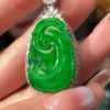 The image displays a beautifully crafted Jadeite Sun Green Ruyi Buckle pendant, held between fingers. The pendant features a rich, bright green jadeite carved in the auspicious shape of a Ruyi, symbolizing good luck and wishes. It's outlined with sparkling stones in a silver setting, with an additional small jadeite stone at the top, creating a luxurious and harmonious piece of jewelry.