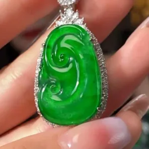 The image displays a beautifully crafted Jadeite Sun Green Ruyi Buckle pendant, held between fingers. The pendant features a rich, bright green jadeite carved in the auspicious shape of a Ruyi, symbolizing good luck and wishes. It's outlined with sparkling stones in a silver setting, with an additional small jadeite stone at the top, creating a luxurious and harmonious piece of jewelry.