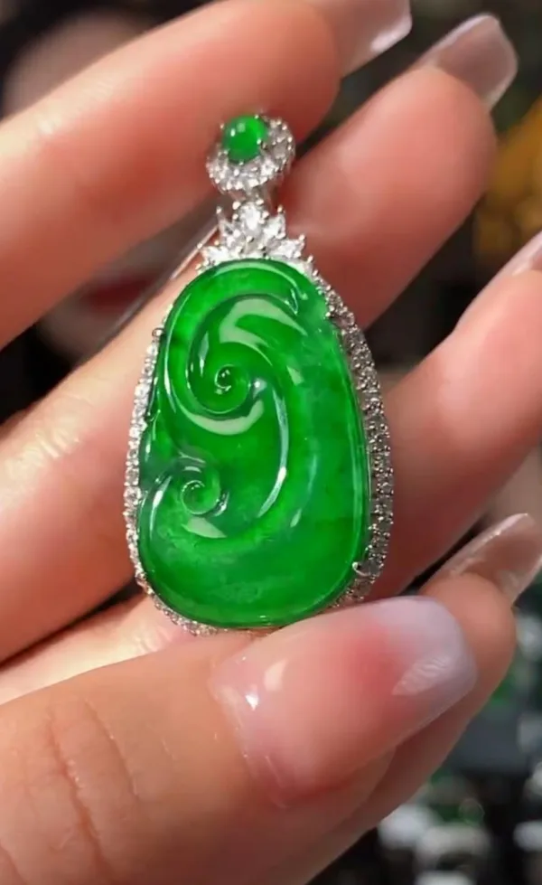 The image displays a beautifully crafted Jadeite Sun Green Ruyi Buckle pendant, held between fingers. The pendant features a rich, bright green jadeite carved in the auspicious shape of a Ruyi, symbolizing good luck and wishes. It's outlined with sparkling stones in a silver setting, with an additional small jadeite stone at the top, creating a luxurious and harmonious piece of jewelry.