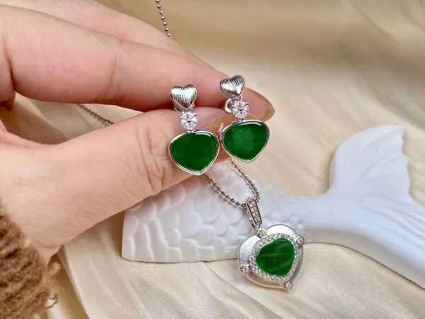 An image showcasing a set of Jade Green Silver Inlay jewelry on a silk and sculpted plaster background. The set includes a heart-shaped pendant with green jade encased in a sparkling silver setting, suspended from a delicate silver chain, and matching heart-shaped stud earrings, each crowned with a small, silver-inlaid heart.