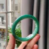 The image shows a hand holding a Full Green Bracelet 61mm Diameter against an urban backdrop seen through a window. The bangle is a rich, even shade of green with a smooth, glossy finish, and it catches the natural light, highlighting subtle variations within the jade. The pure green color speaks of serenity and the natural world, contrasting beautifully with the modern setting in the background.