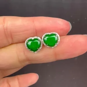 A hand displaying a pair of Ruyi Matching Stud Earrings (Gold Inlaid), featuring heart-shaped vivid green jade set in an ornate gold inlay, encased in a silver outline dotted with small sparkling stones that enhance the jade's natural beauty.