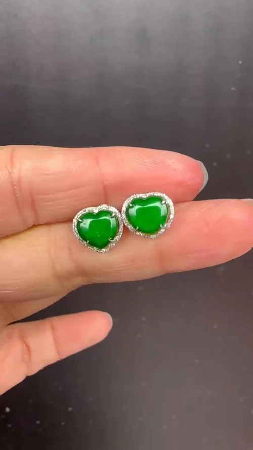 A hand displaying a pair of Ruyi Matching Stud Earrings (Gold Inlaid), featuring heart-shaped vivid green jade set in an ornate gold inlay, encased in a silver outline dotted with small sparkling stones that enhance the jade's natural beauty.