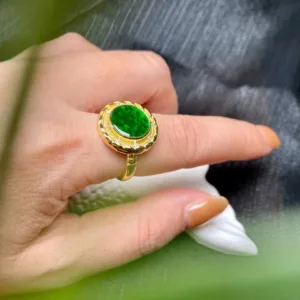 A hand gracefully displaying a "Jade Green Ring with Silver Inlay" featuring a radiant, oval-cut green jade gemstone at its center. The jade is encased in a gold setting, adorned with intricate silver inlay and a halo of smaller sparkling stones that catch the light, lending the piece an opulent and sophisticated charm.