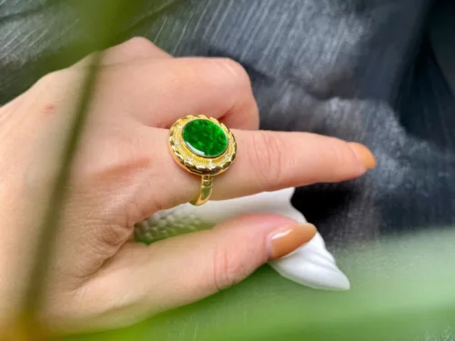 A hand gracefully displaying a "Jade Green Ring with Silver Inlay" featuring a radiant, oval-cut green jade gemstone at its center. The jade is encased in a gold setting, adorned with intricate silver inlay and a halo of smaller sparkling stones that catch the light, lending the piece an opulent and sophisticated charm.