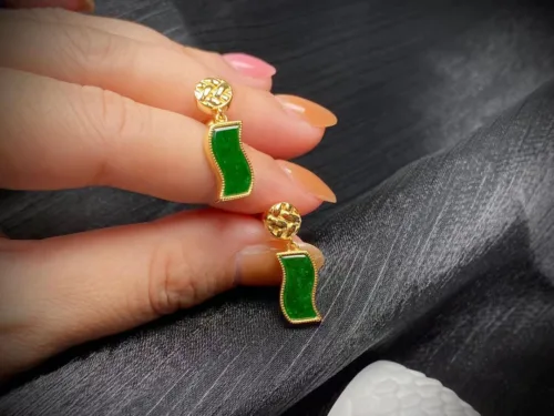 A close-up photo of a hand displaying a pair of Jade Green Earrings with Silver Inlay, highlighting the rich green color of the jade set against the brilliant silver outline. The earrings feature a unique shape, reminiscent of a banner or flag, and are topped with a golden, textured stud, creating an elegant contrast and adding a sophisticated touch to the overall design.